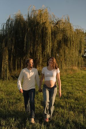 Happy couple walking together in grassy field near weeping willow tree at golden hour.