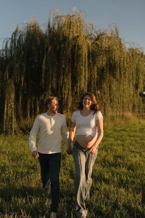 Happy couple walking hand-in-hand in field at golden hour, pregnant woman in white top holding baby bump.