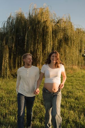 Happy pregnant couple holding hands outdoors near a willow tree in golden hour sunlight, woman showing baby bump.
