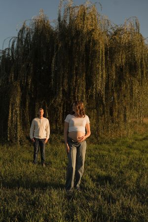 Couple outdoors at sunset near weeping willow, pregnant woman in white top holding baby bump with partner behind.