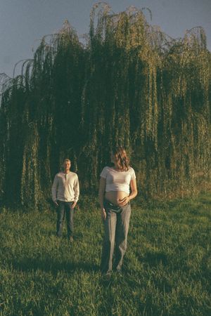 Pregnant woman cradling baby bump standing near weeping willow tree with partner in background at dusk.