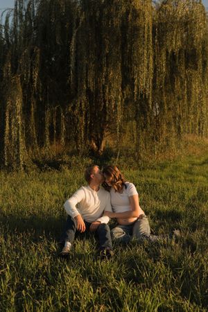 Couple sitting on grass kissing under a weeping willow tree at golden hour sunset.