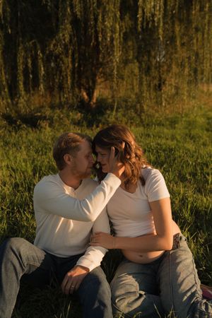 Couple sitting in grass under willow tree at golden hour, man tenderly touching pregnant woman's face.