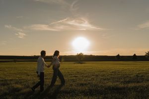 Couple holding hands walking across a lush green field during a stunning sunset with glowing sky.