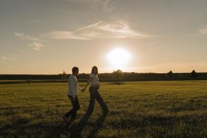 Silhouette of pregnant couple walking hand-in-hand across green field during stunning golden sunset.