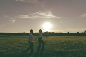 Couple holding hands in a sunlit green field at sunset, silhouetted against a glowing sky.