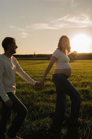 Pregnant couple holding hands in a sunlit green field at golden hour, woman smiling with baby bump visible.