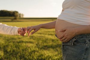 Close-up of couple holding hands outdoors in green field, pregnant woman in white top cradling baby bump.