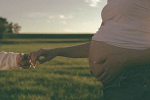 Intimate close-up of couple holding hands in green field, pregnant woman in jeans gently holding baby bump.