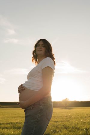 Pregnant woman in white tee and jeans cradling her baby bump in a sunlit green field at golden hour.