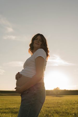 Pregnant woman in white tee and jeans cradling her belly at golden hour in a green field.