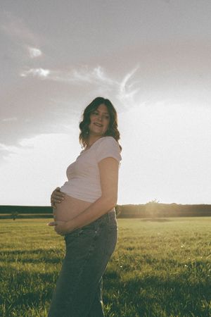 Pregnant woman in white tee and jeans cradling baby bump in a sunlit green field at golden hour.