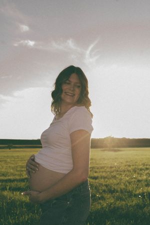 Smiling pregnant woman in white t-shirt cradling baby bump outdoors in green field at golden sunset.