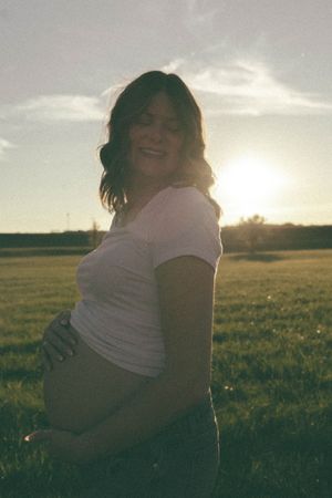 Pregnant woman in white t-shirt smiling at sunset in a green field, golden backlight creating a warm glow.