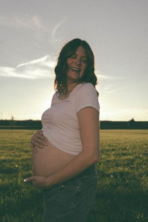 Happy pregnant woman laughing while holding baby bump in sunlit green field during golden hour.