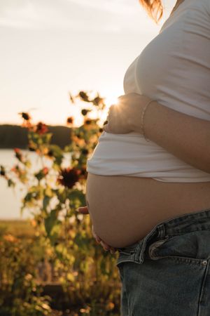 Close-up of pregnant woman in white top and jeans holding bare baby bump near sunflowers at sunset.