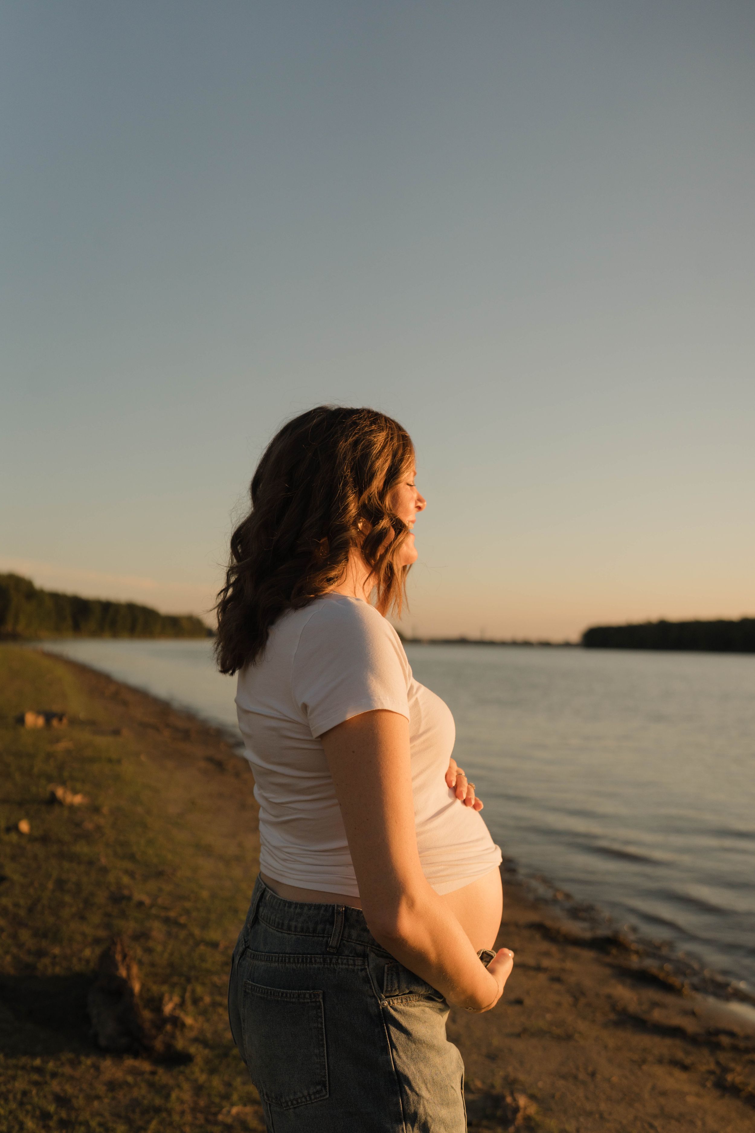 Pregnant woman in white top and jeans cradling her baby bump by a serene lakeside at golden hour sunset.