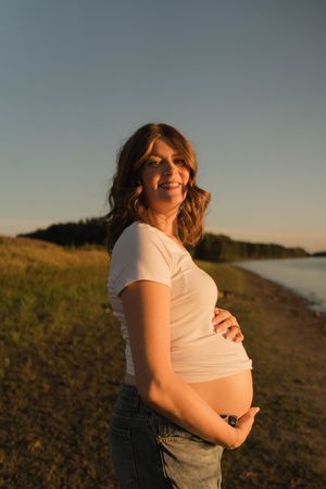 Smiling pregnant woman in white top and jeans cradling her baby bump outdoors at golden hour near a lake.