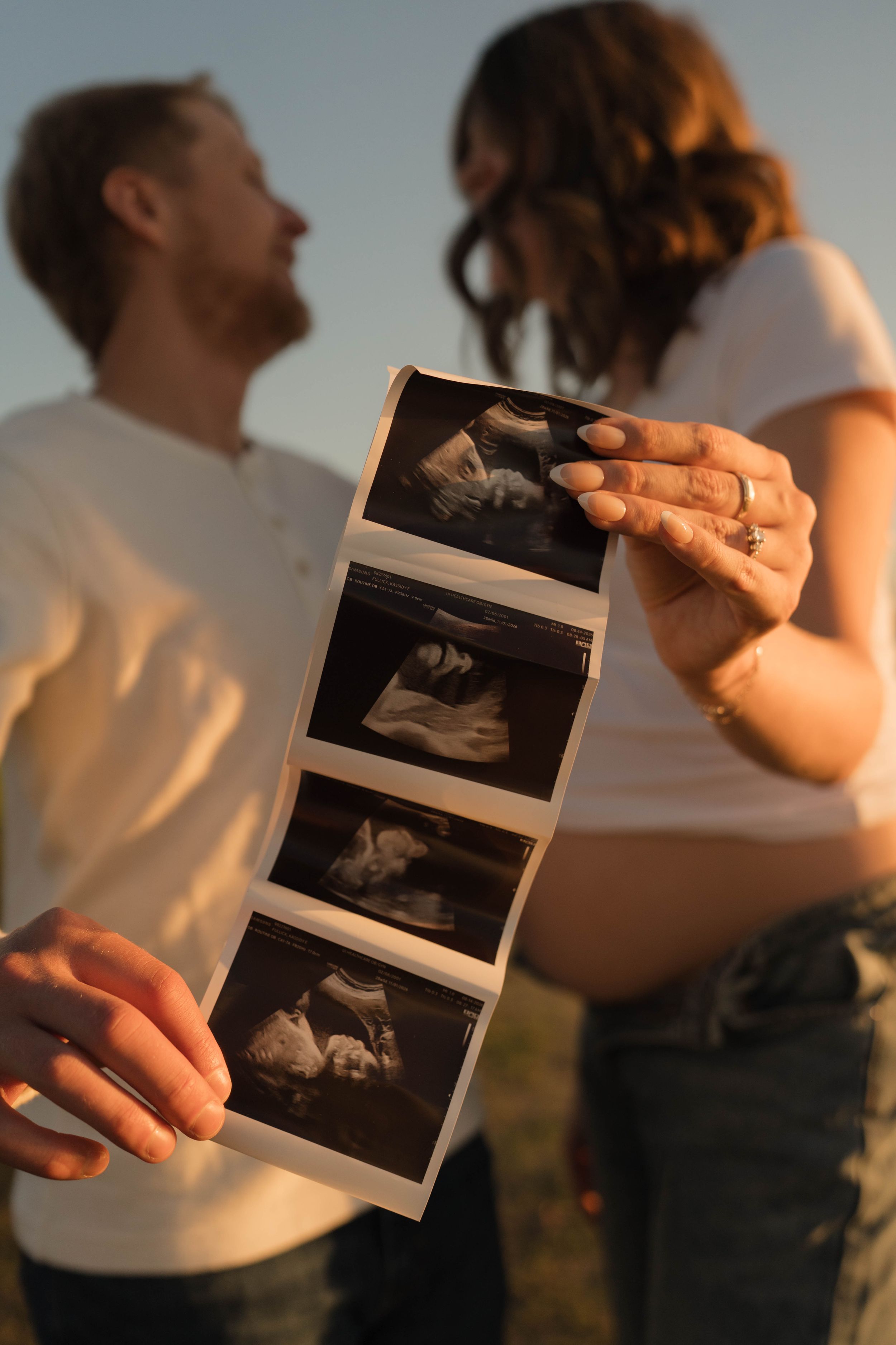 Couple holding ultrasound photos outdoors at sunset, pregnant woman and partner sharing joyful moment.