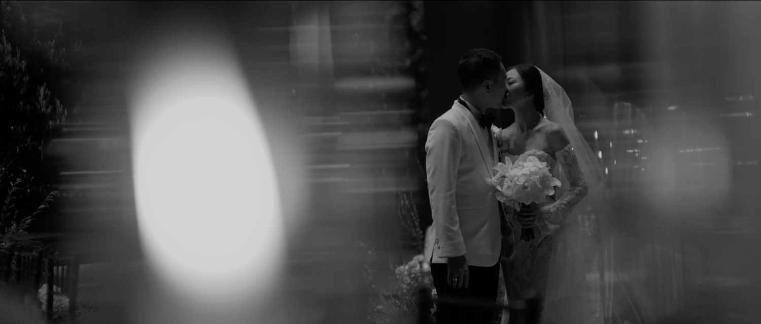 Black and white wedding photo of a couple sharing an intimate moment, bride holding bouquet with soft bokeh lighting.