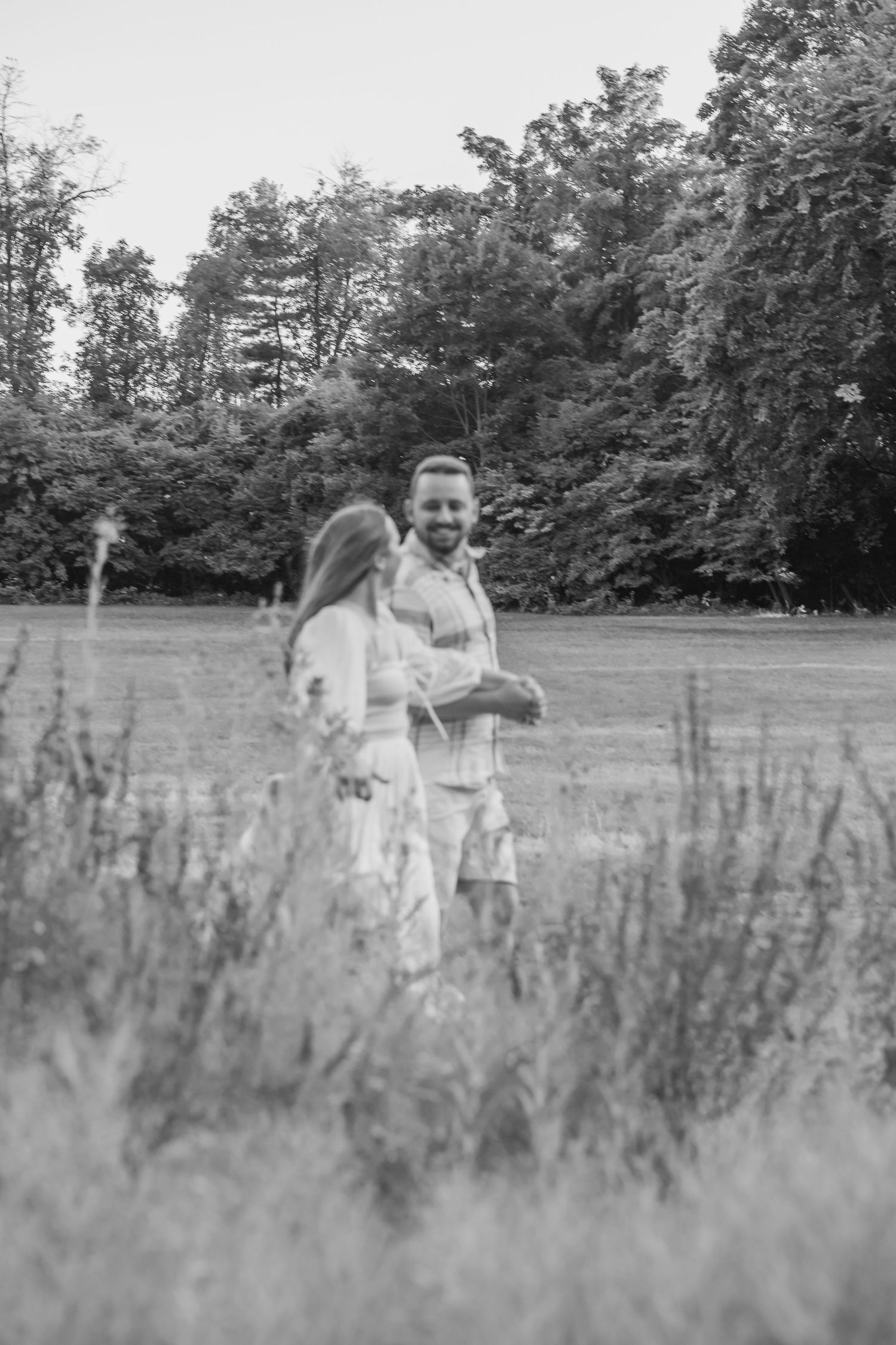 Black and white photo of a smiling couple standing in a meadow with tall grass and trees in the background.