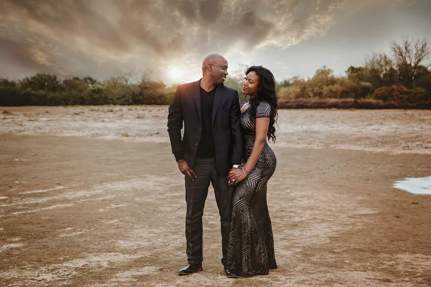 A couple in dark formal outfits embraces on a dry cracked lakebed at sunset beneath a dramatic cloudy sky.