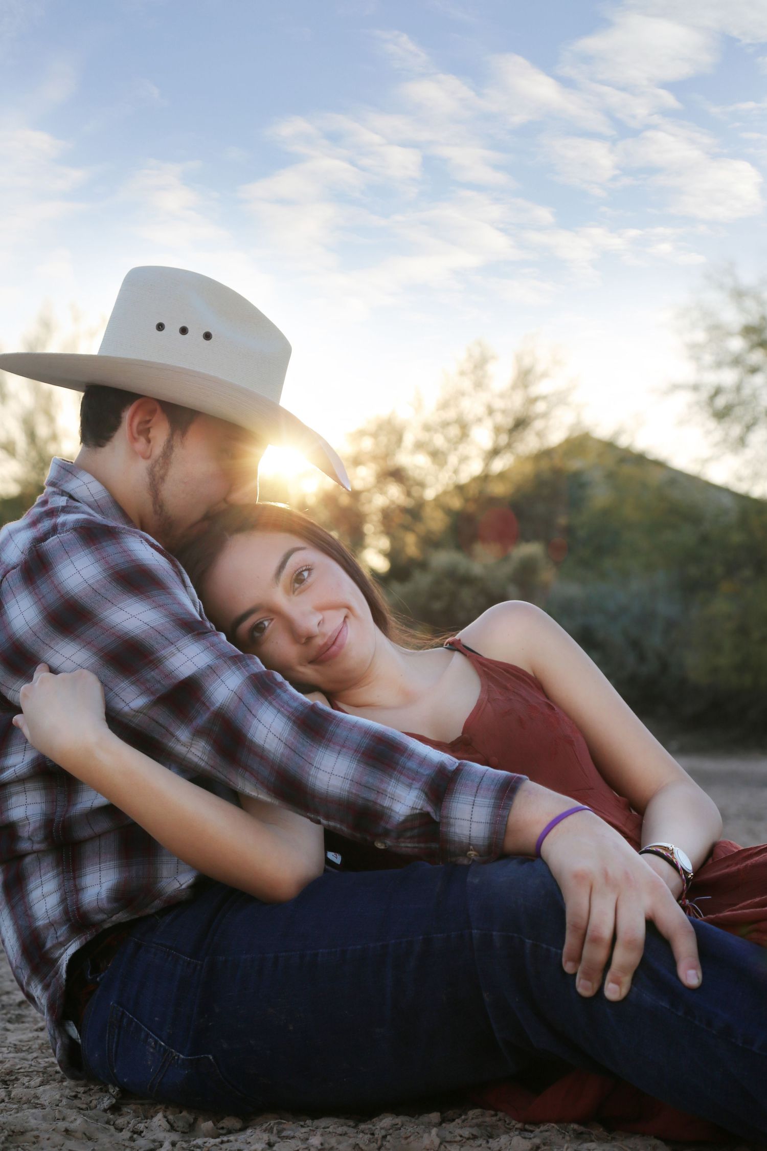 A man in a cowboy hat kisses the forehead of a smiling woman as they sit close together outdoors at golden hour.