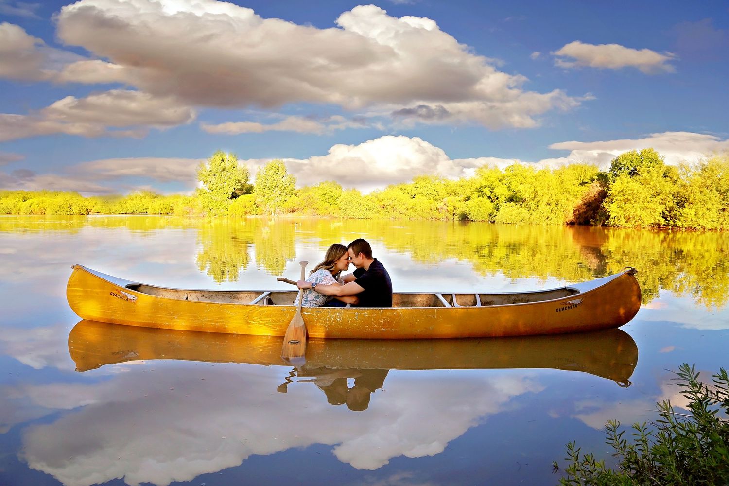 A couple shares a kiss in a yellow canoe on a calm lake with golden trees and clouds reflected in the water.