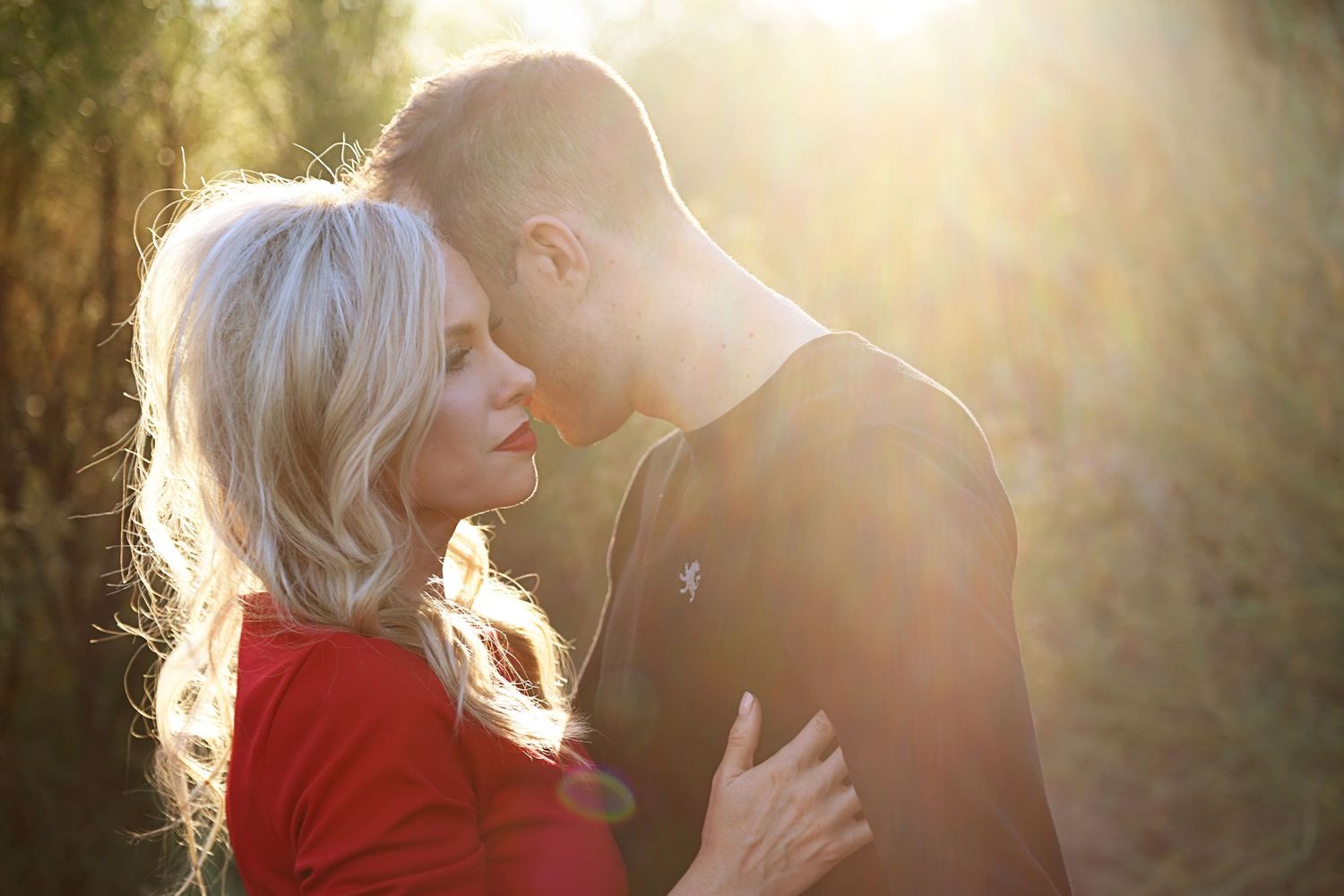 A blonde woman in a red top and a man embrace in warm, glowing sunlight surrounded by trees.
