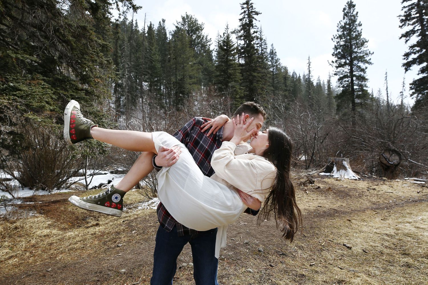 A man in a plaid shirt cradles and kisses a woman in a white dress on a forest trail with snow and pine trees.