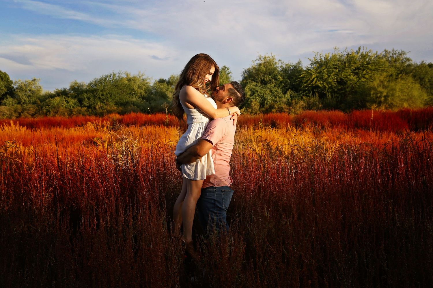 A man lifts a woman in a white dress in a loving embrace in a vivid field of red and orange grasses.