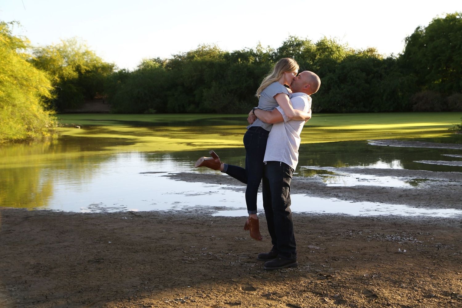 Couple embracing as the groom lifts the bride beside a calm pond with green trees in the background.