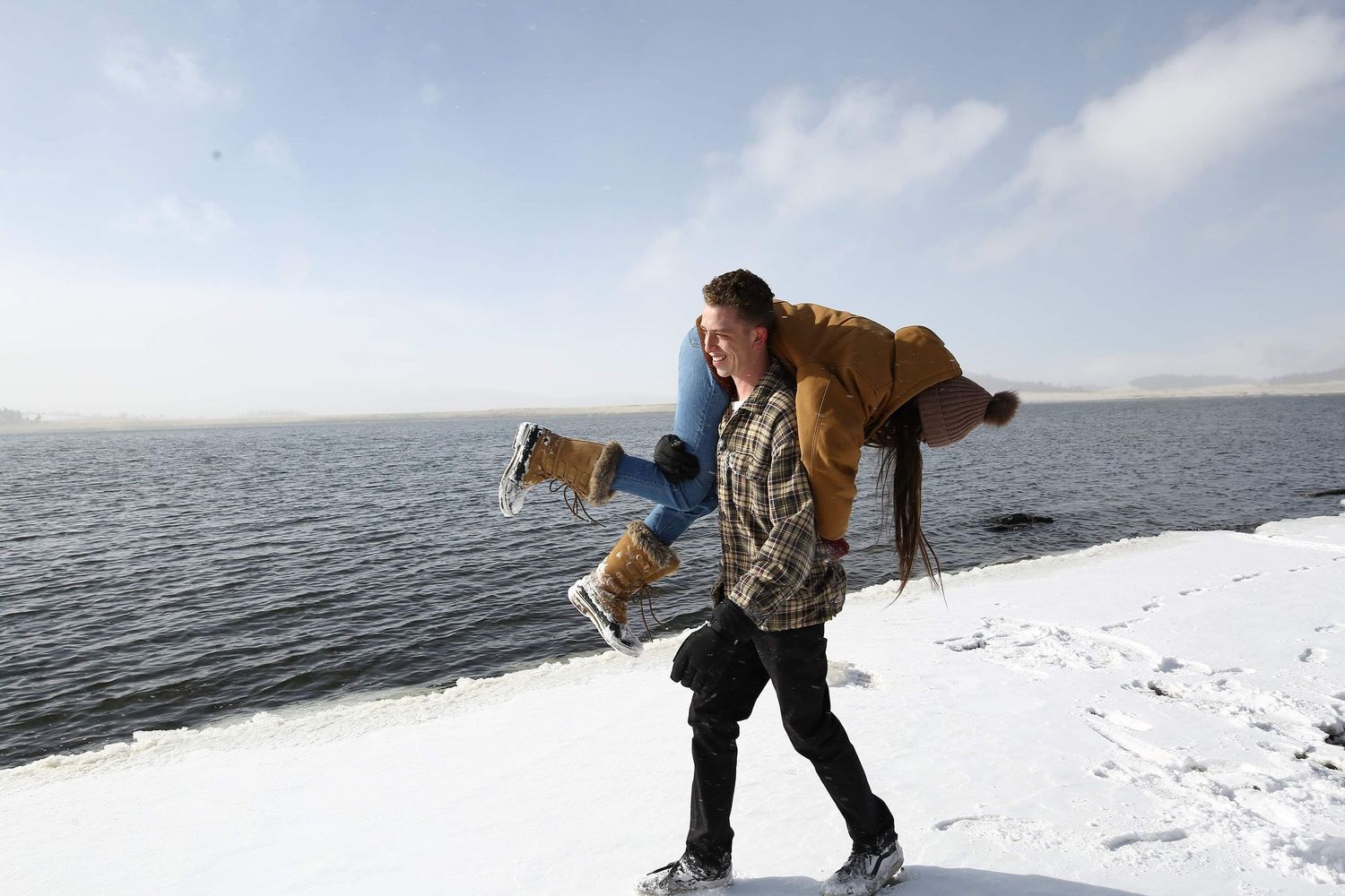A man in winter clothes carries a woman in boots on his back along a snowy shore beside a calm lake.