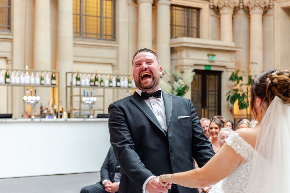 Groom laughing joyfully during wedding ceremony while holding brides hands in elegant venue.