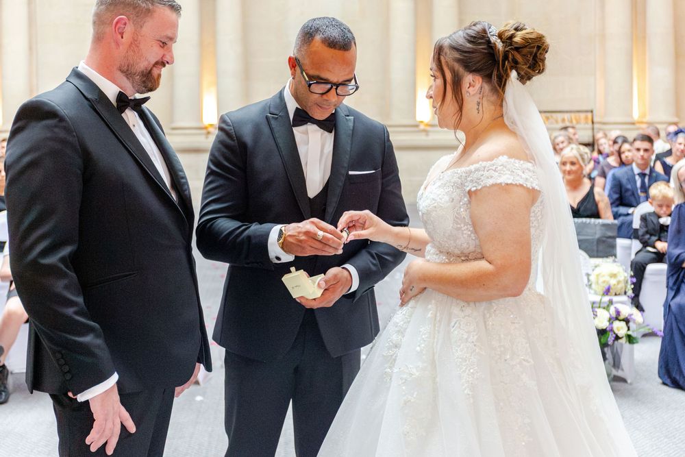 Groom places wedding ring on brides finger during elegant ceremony, officiant watching, guests seated in ornate venue.