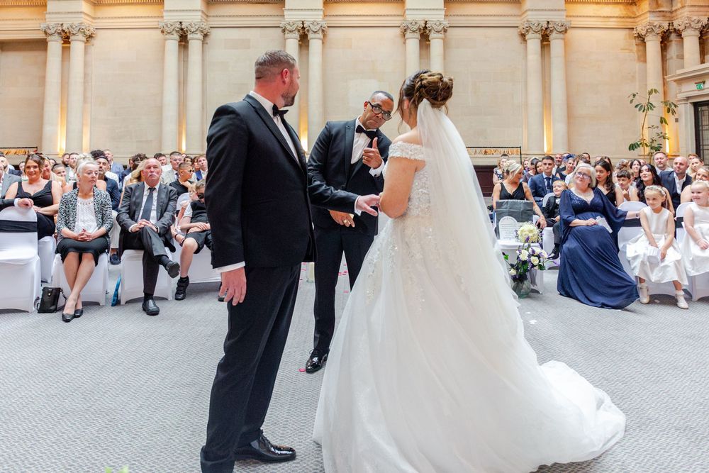 Bride and groom exchange vows at elegant indoor wedding ceremony with guests seated in background.