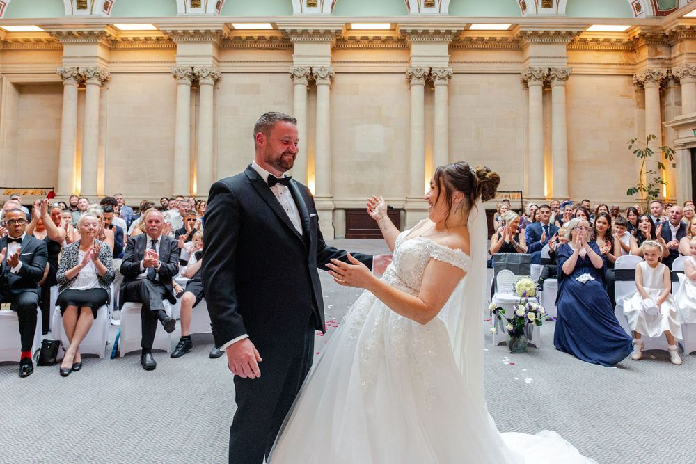 Bride reads vows to groom in black tuxedo during elegant wedding ceremony with guests applauding in grand venue.