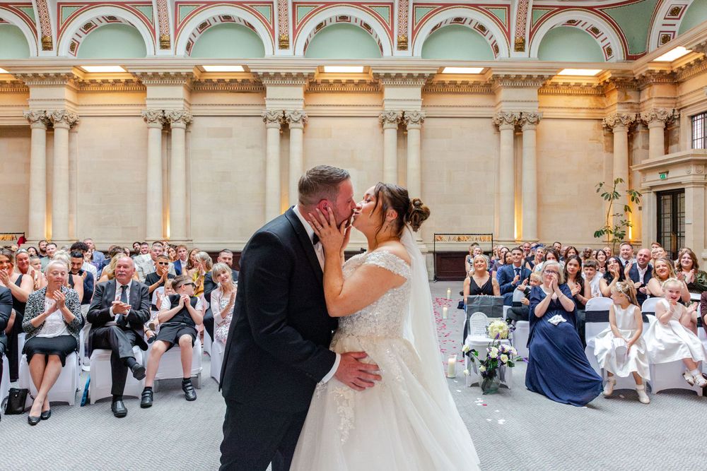 Bride and groom share first kiss at altar during wedding ceremony with applauding guests in ornate ballroom.