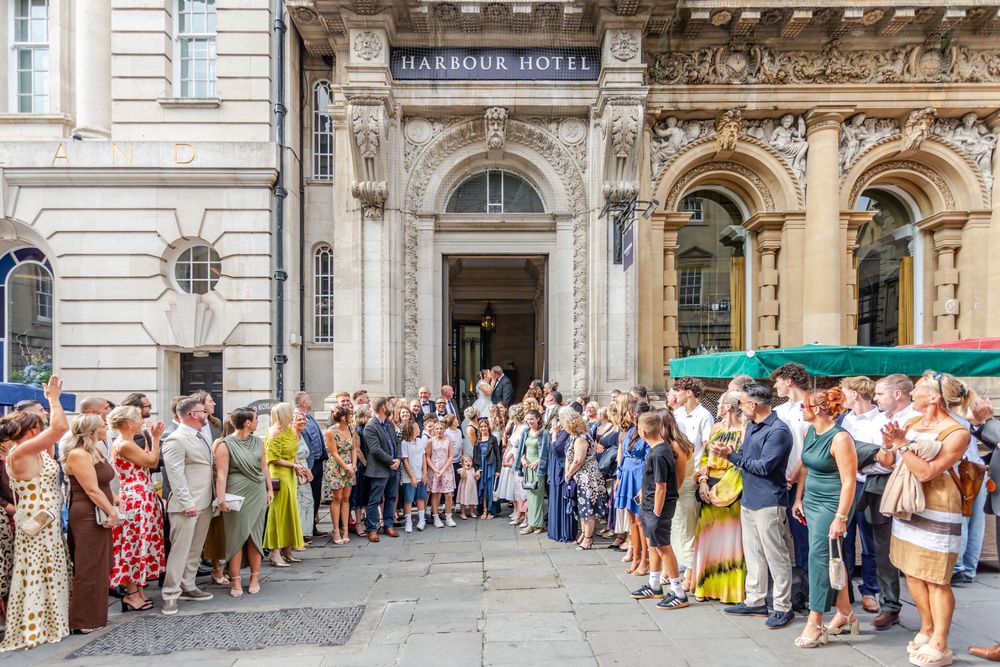 Crowd of elegantly dressed guests gathered outside the historic Harbour Hotel entrance on a sunny day.