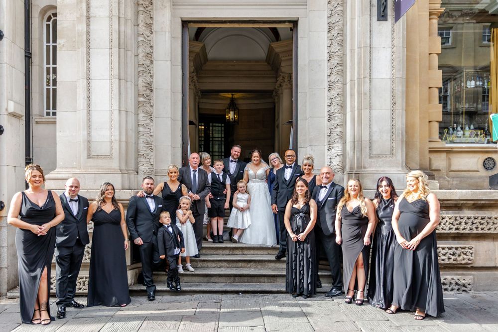 Wedding party posing on steps of elegant stone building, bride and groom surrounded by guests in black formal attire.