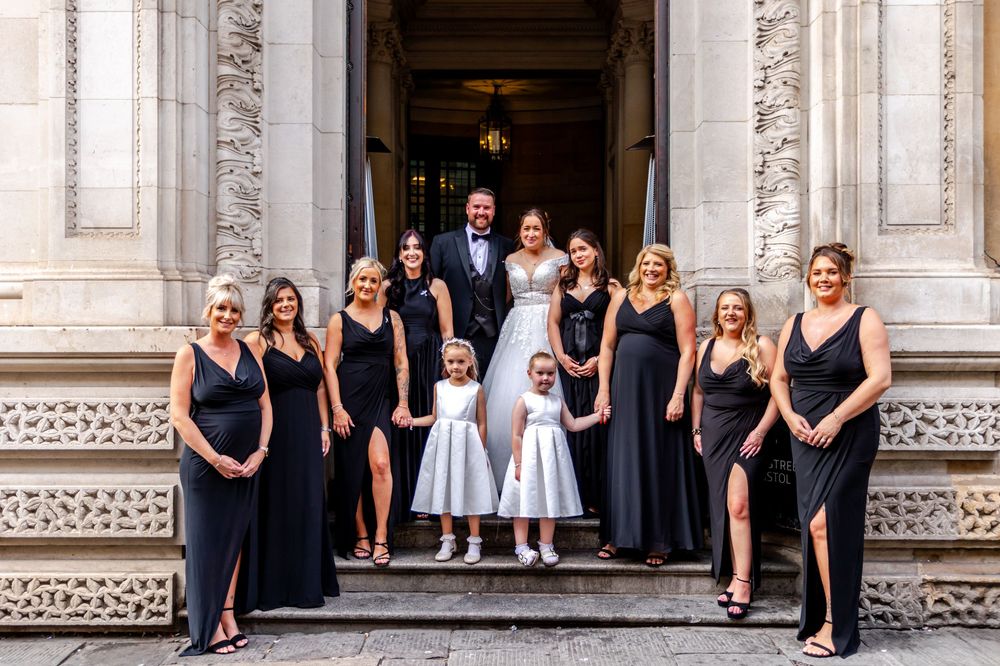 Wedding party poses on stone steps, bride and groom flanked by bridesmaids in black gowns and two flower girls in white.