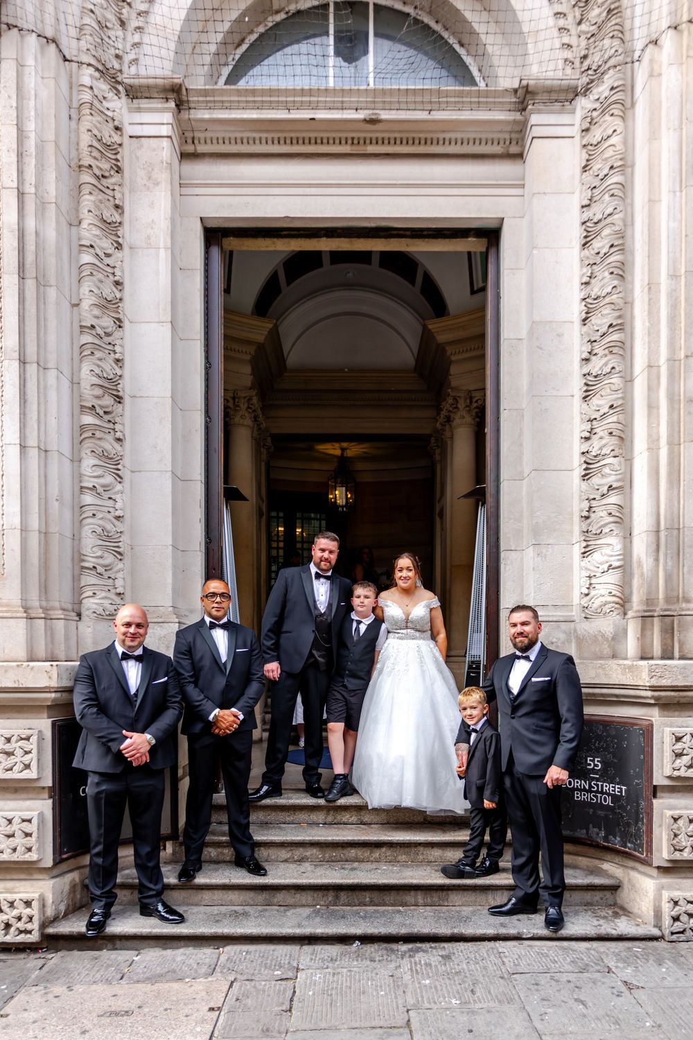 Wedding party posing on steps of elegant stone building, bride in white gown with groom and groomsmen in tuxedos.