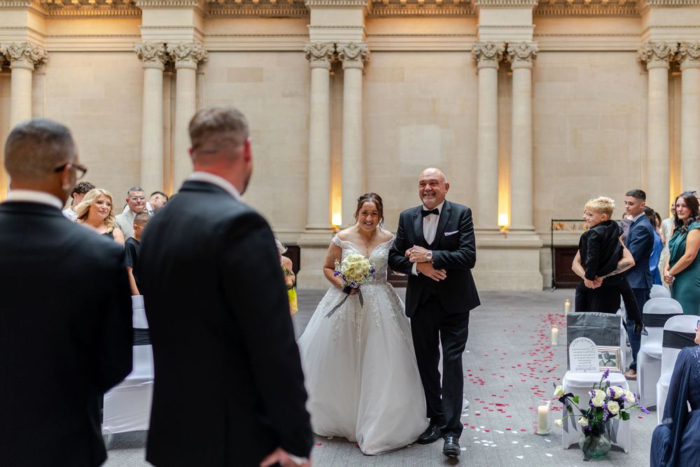 Bride walks down the aisle with her father in an elegant wedding ceremony held in a grand classical venue.