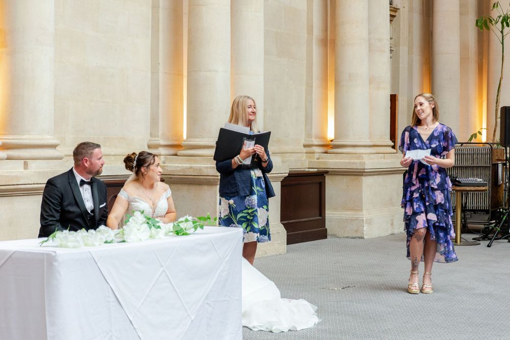Bride and groom seated at wedding table while officiant and guest speaker address them in elegant outdoor venue.