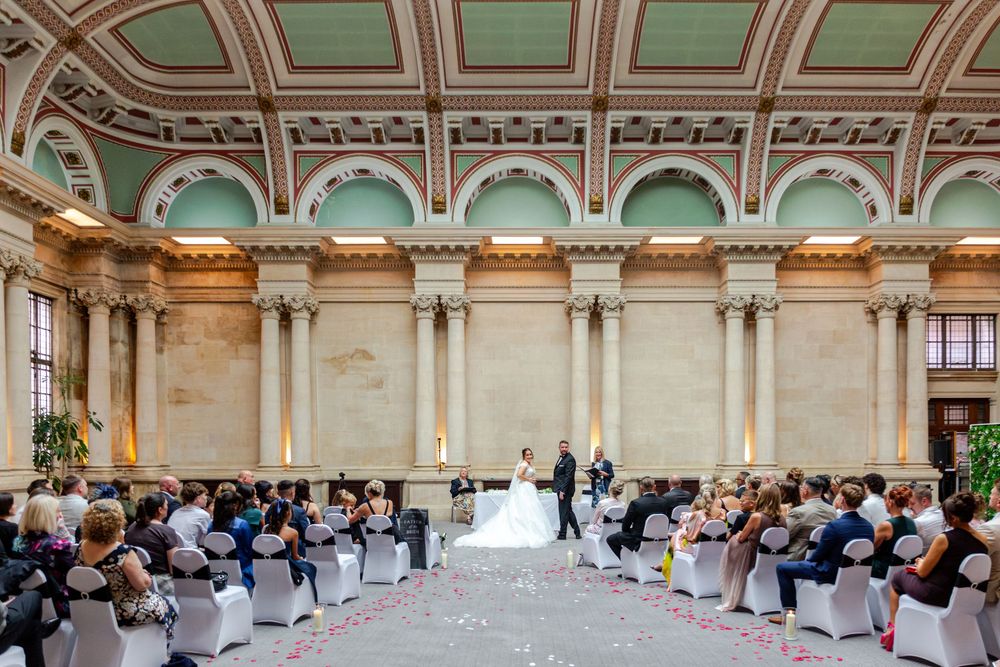 Elegant wedding ceremony in a grand neoclassical hall with tall columns, ornate ceiling, and guests seated along a rose petal aisle.