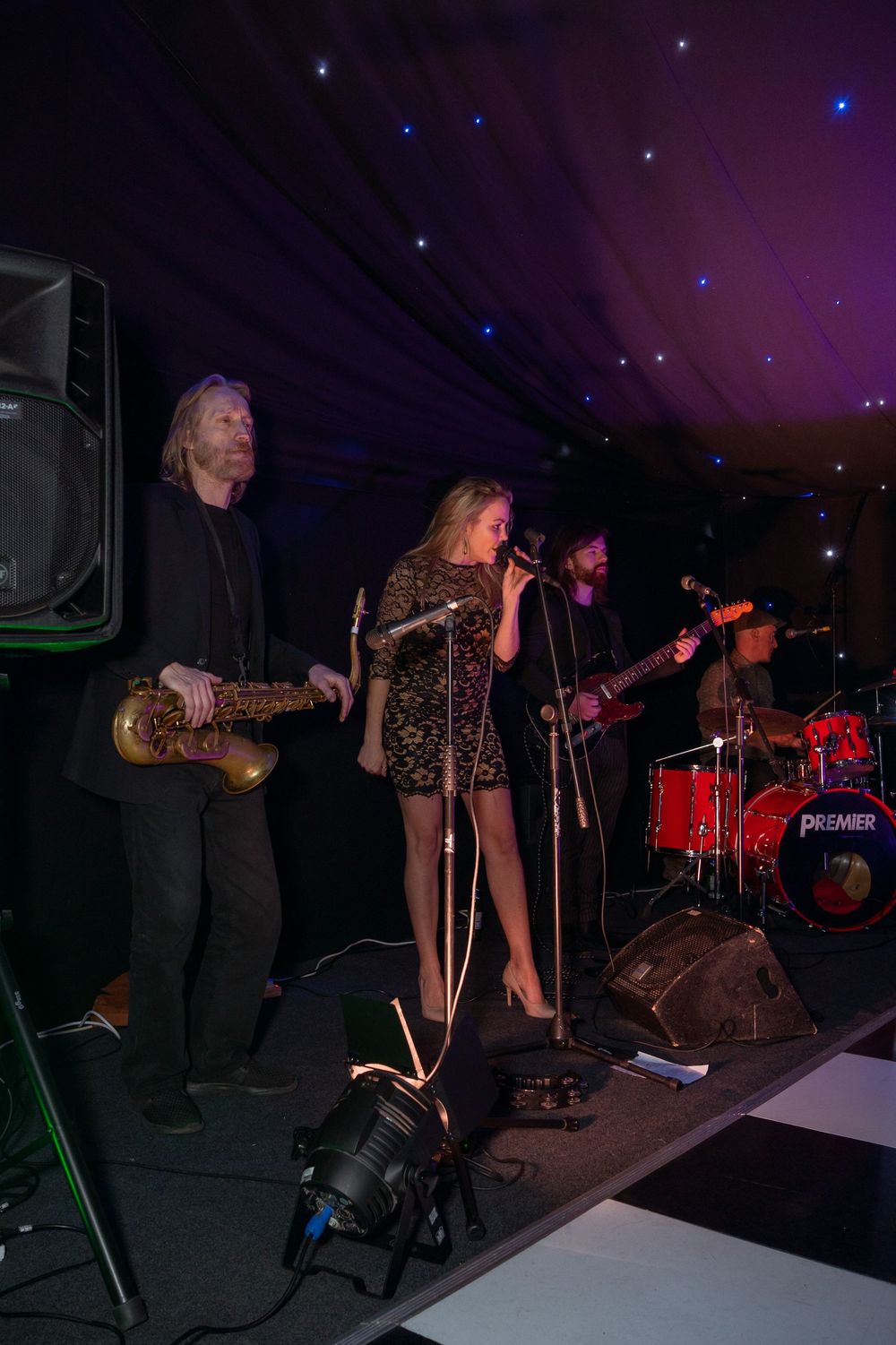 Live band performing on stage with saxophonist, female vocalist, guitarist, and drummer in a marquee venue.