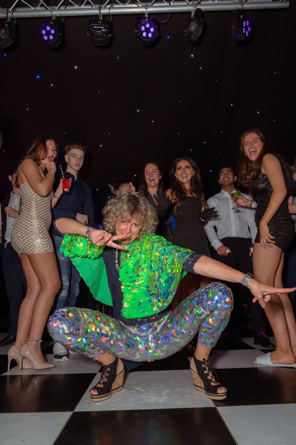 Person in green sequin top and holographic pants dancing at a party with crowd cheering behind them.