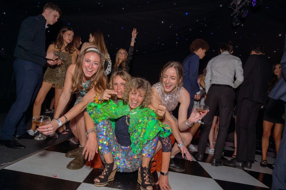 Three young women in sequin dresses pose and dance together on a checkered dance floor at a festive party event.