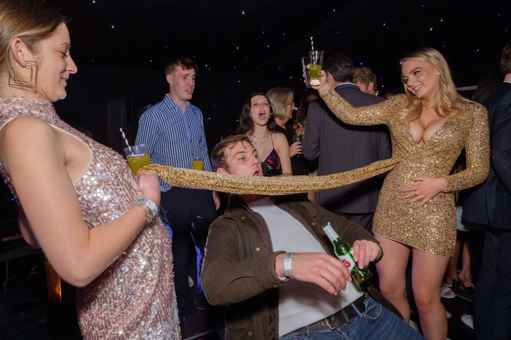 Young adults in formal attire enjoy a lively party, playing a limbo game with drinks in hand at a nightclub.