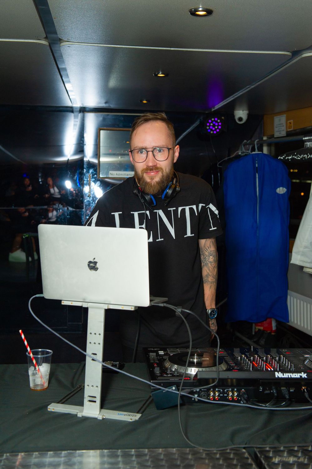 Bearded male DJ with glasses using Apple MacBook laptop and Numark mixer at a nightclub event.